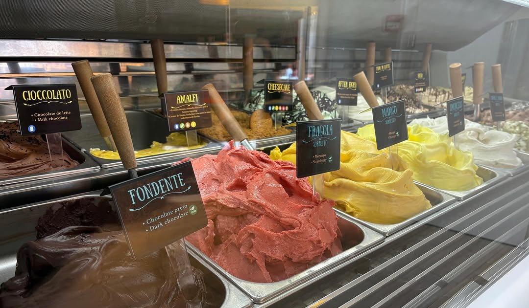 Vitrine de gelados com tinas de gelado castanho, vermelho e amarelo, com espátulas e etiquetas com os nomes dos sabores